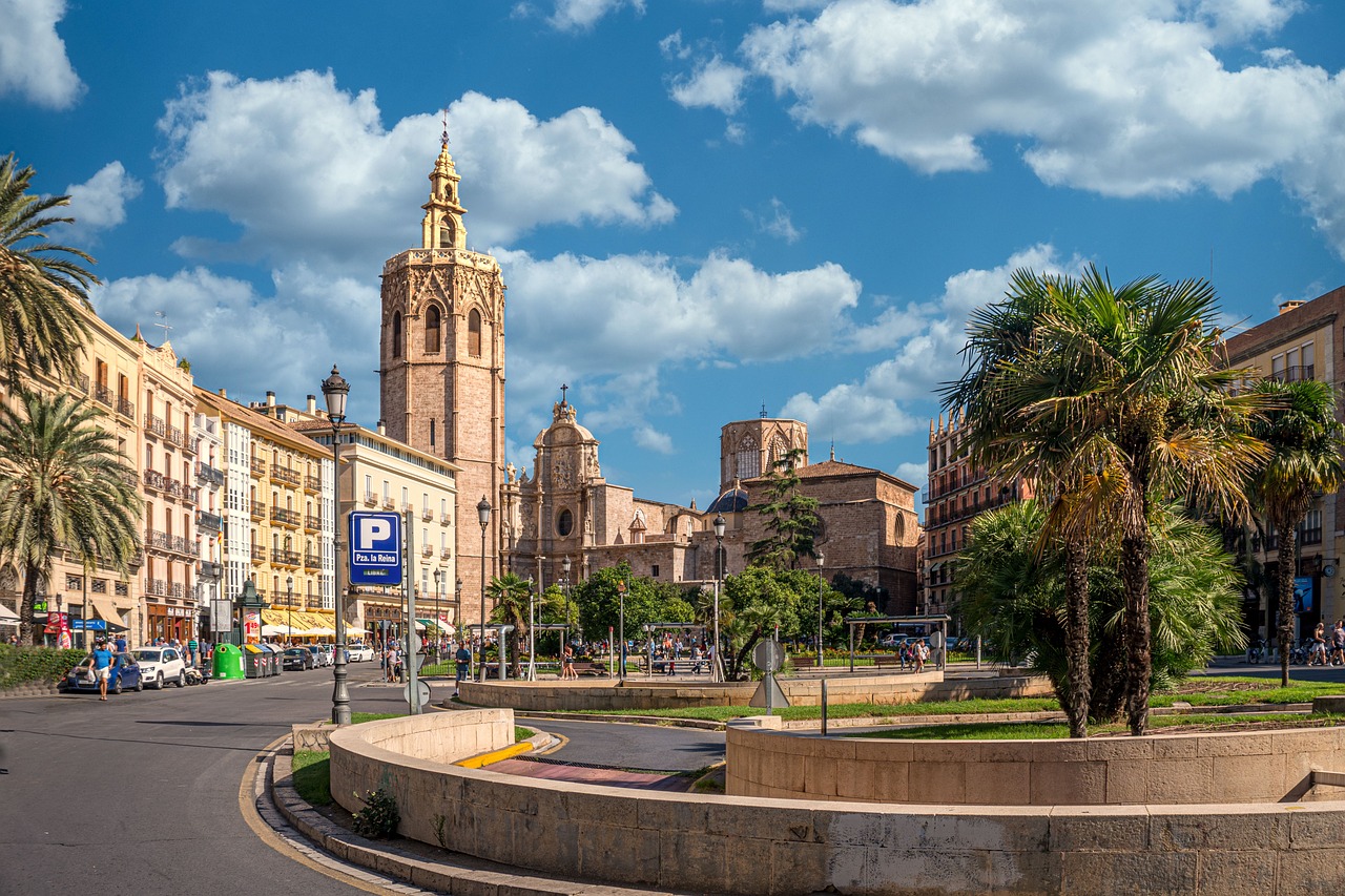 Valencia travel guide: View of Valencia Cathedral and El Miguelete Tower with palm trees and historic buildings in the foreground under a blue sky with clouds.