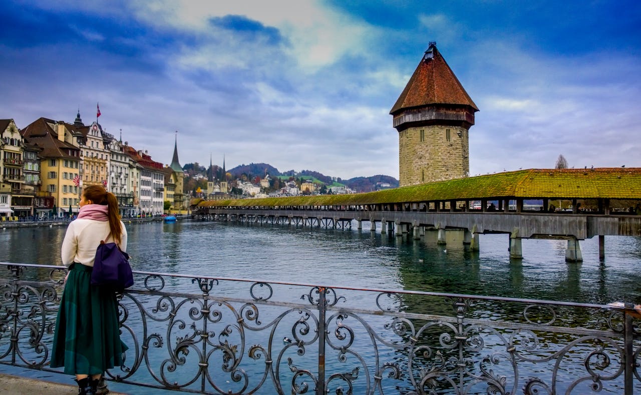 A woman gazes at the historic Chapel Bridge (Kapellbrücke) and its iconic water tower in Lucerne, Switzerland, with the city's scenic lake and medieval architecture in the background.